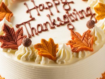 A round cake with "Happy Thanksgiving" written on top in brown icing. Decorated with autumn-colored sugar leaves and small acorn shapes around the edge. The sides have textured frosting and orange sprinkles at the base.