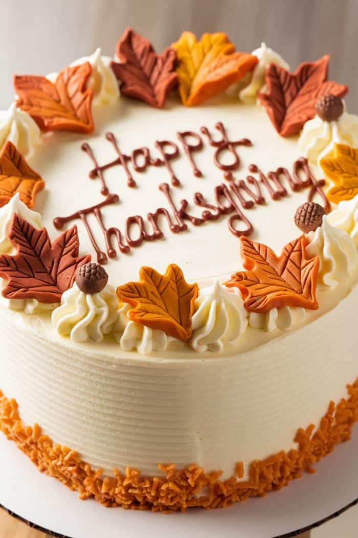 A round cake with "Happy Thanksgiving" written on top in brown icing. Decorated with autumn-colored sugar leaves and small acorn shapes around the edge. The sides have textured frosting and orange sprinkles at the base.