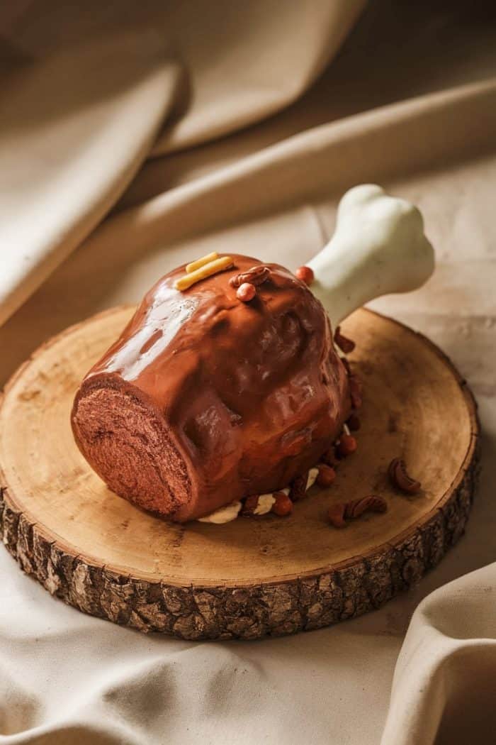 A realistic cake shaped like a large turkey leg, complete with a bone made from fondant, sits on a wooden slab. The surface is smooth and glazed, resembling roasted meat, surrounded by nuts for decoration.