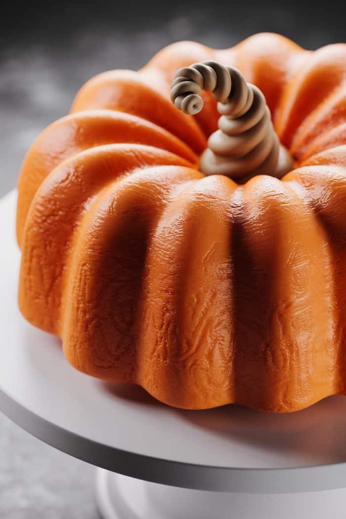 A cake designed to look like a pumpkin with an orange, ridged surface and a swirled stem sits on a white stand, set against a soft-focus gray background.