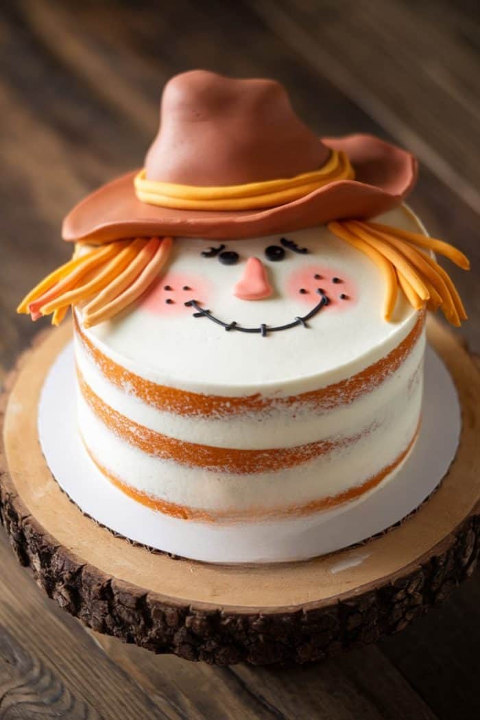 A round cake decorated to look like a scarecrow with a fondant cowboy hat, orange fondant hair, and a smiling face with rosy cheeks. The cake has a semi-naked frosting style and is placed on a wooden platter.
