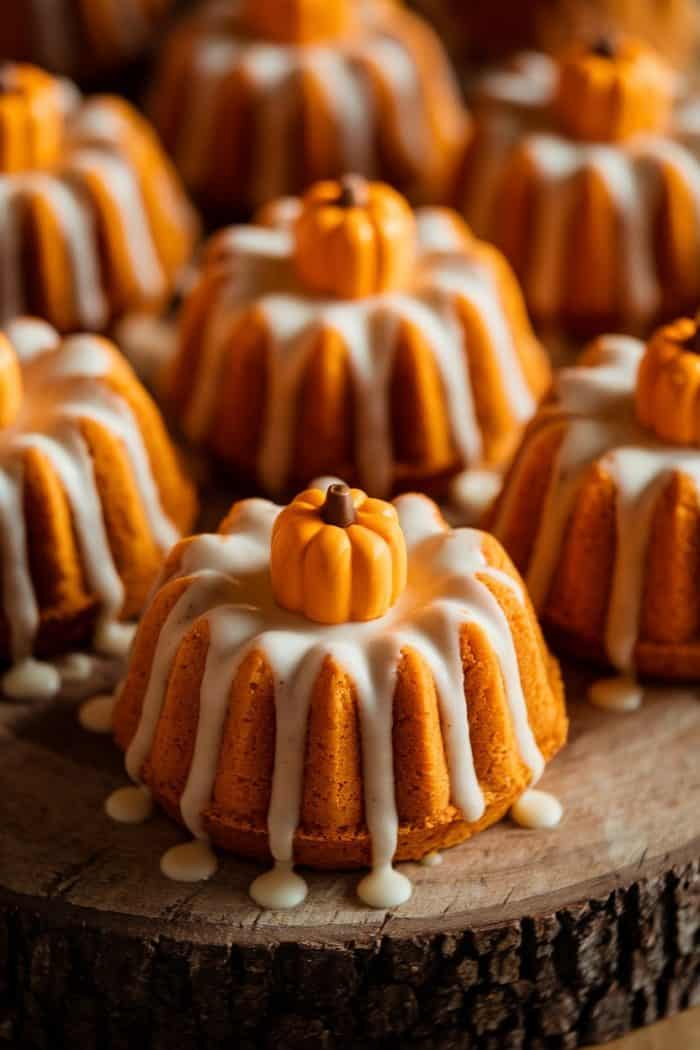 Miniature bundt cakes topped with white icing and decorated with small pumpkin-shaped candies are displayed on a rustic wooden surface. The warm colors and autumn-themed decorations evoke a seasonal, festive atmosphere.