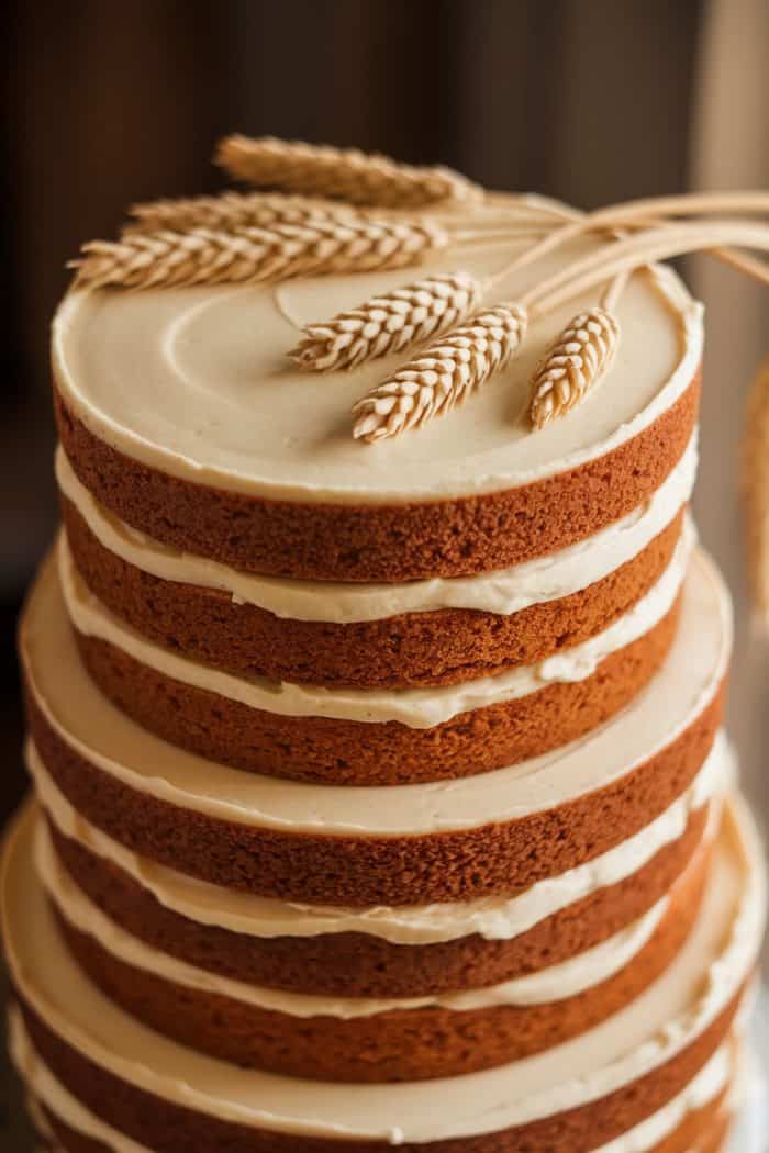 A tiered naked cake topped with sheaves of wheat. The cake has visible layers of cream between the golden brown sponge. The rustic decoration gives it a natural, earthy appearance.