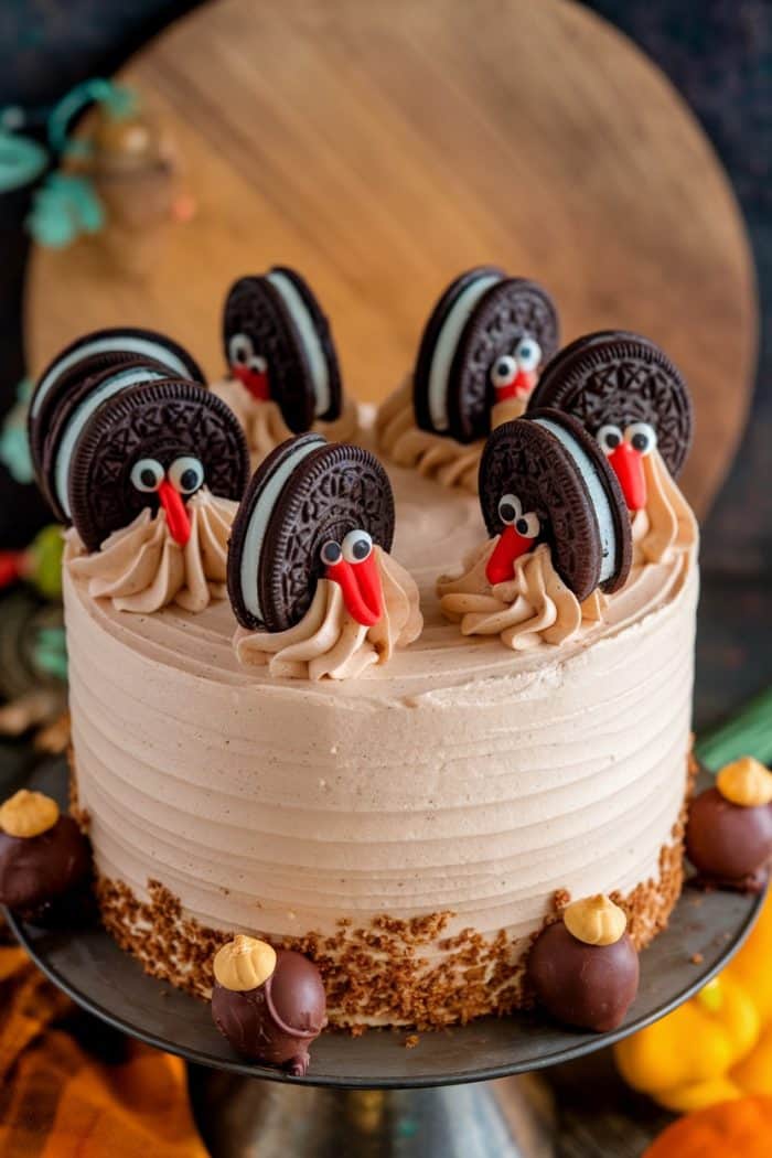A festive cake decorated with cookies and cream turkeys. Each turkey is made with a cookie for the body and candy eyes and a red candy for the beak. The cake features swirled frosting and chocolate truffles around the base.