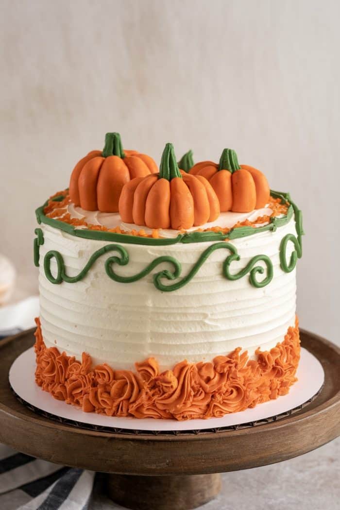 A festive cake decorated with white and orange frosting, topped with three orange pumpkin shapes and green icing accents. The cake sits on a wooden stand, with a textured and layered design.