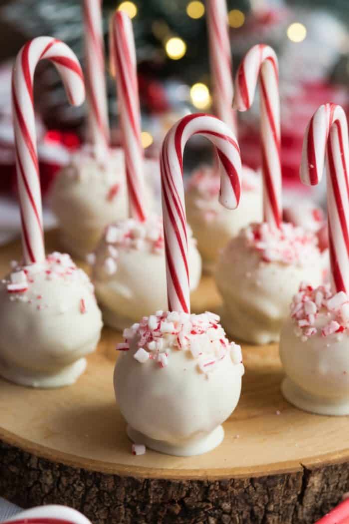 Festive cake pops coated in white chocolate are topped with crushed peppermint and decorated with candy canes. They are arranged on a round wooden platter with blurred holiday lights in the background.
