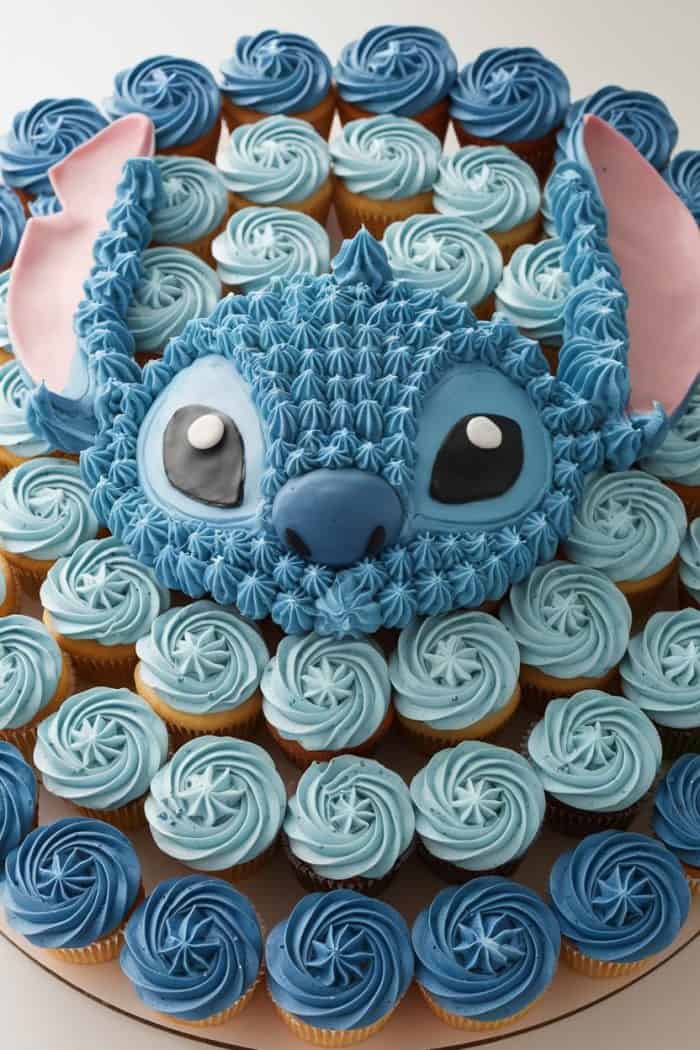A cake ensemble featuring a large, detailed blue character face with big eyes and pink ears atop a circular arrangement of cupcakes. The cupcakes are frosted in varying shades of blue and white, creating a cohesive design.