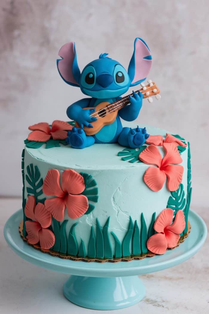 A turquoise cake adorned with a blue alien figure playing a guitar sits on a matching cake stand. The cake is decorated with pink hibiscus flowers and green leaves, creating a tropical theme.