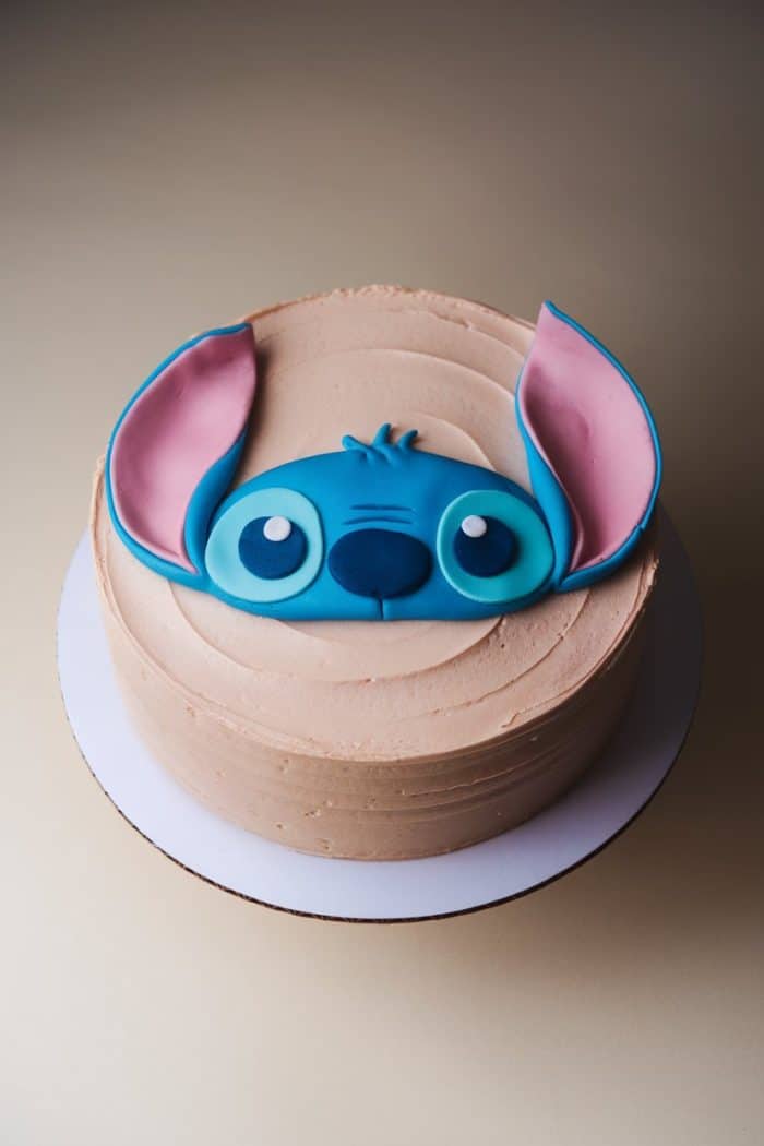 A round cake with tan frosting features a blue icing decoration resembling a character with large ears, big eyes, and a small nose on the top. The cake is displayed on a white cake board.