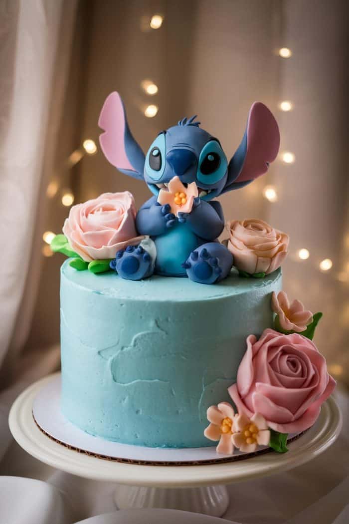 A light blue cake with a fondant figure of a blue, cartoon-like creature holding a star, surrounded by pink and peach flowers. The cake is displayed on a white stand, with fairy lights in the blurred background.