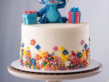 A white cake adorned with colorful flowers and beads. Atop sits a blue alien figure holding balloons, surrounded by gift boxes. The cake rests on a beige stand with decorative paper fans around it.