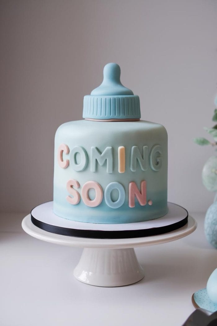 A baby-themed cake shaped like a blue baby bottle, with the words "COMING SOON" in pastel colors. It sits on a white cake stand with a black ribbon.
