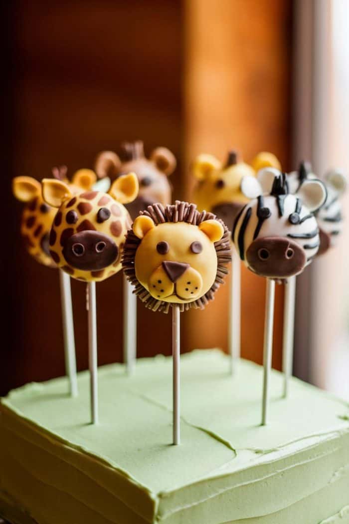 Colorful animal cake pops, featuring giraffes, a lion, and zebras, are arranged in a group on top of a green cake base. The pops are intricately detailed with chocolate accents and attached to white sticks.