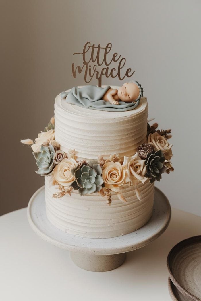 Two-tiered white cake with a "little miracle" topper, adorned with beige roses and succulents. A sleeping baby figure rests on top, covered with a small gray blanket. The cake is on a white stand, with a simple gray background.