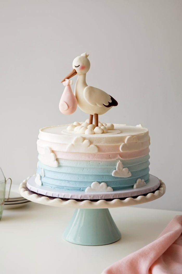 A pastel-colored cake with a stork topper carrying a pink bundle. The cake has layers in blue, pink, and white with cloud decorations. It's displayed on a light blue cake stand.