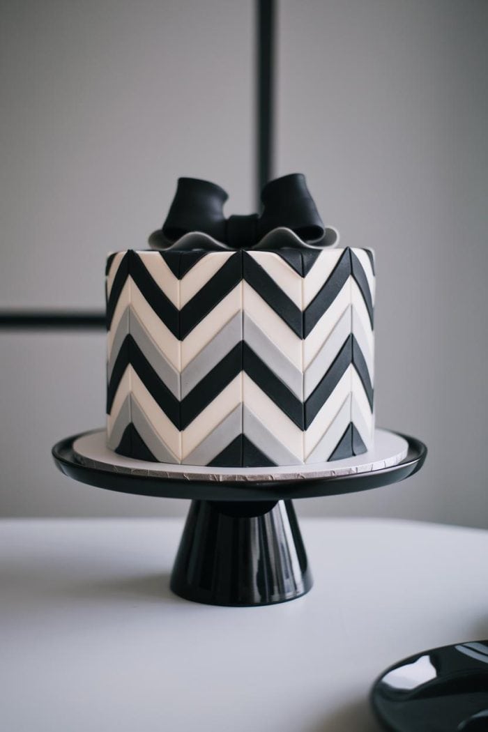A round cake with a black and white chevron pattern on a black pedestal stand, topped with black ribbon-like decorations, set against a minimalist background.