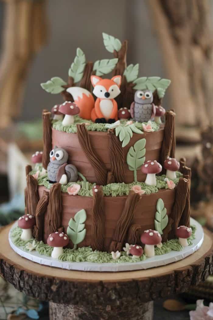 A three-tiered woodland-themed cake decorated with brown frosting resembling tree bark. It features fondant animals, including a fox, owls, and a squirrel, as well as mushrooms and green leaves, all on a wooden cake stand.