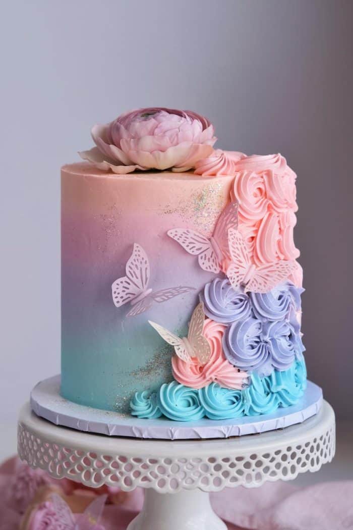 A pastel-colored cake with ombre shades of pink, purple, and blue. Decorated with frosting rosettes, cutout butterfly shapes, and topped with a large peony flower. It sits on a white cake stand with lace-like edges.