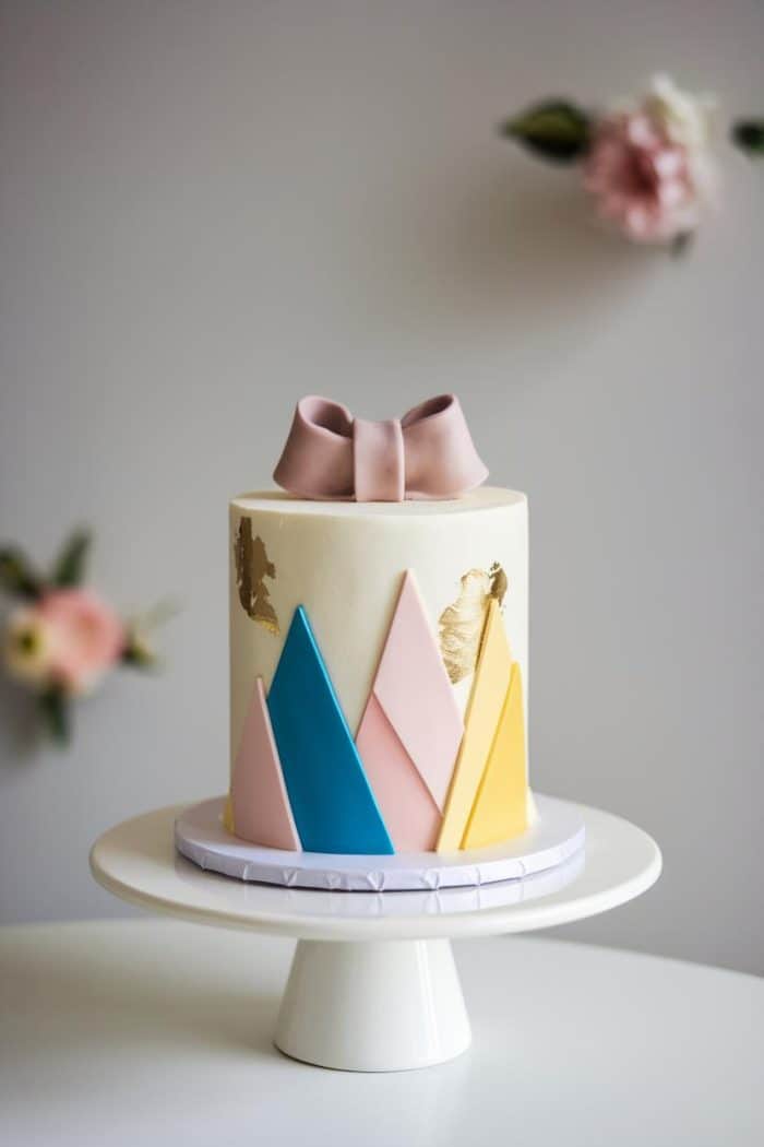 A tall, elegant cake with a smooth cream-colored frosting, decorated with blue, pink, and yellow angular shapes and gold leaf accents. A pink bow sits on top. It's placed on a white cake stand with blurred flowers in the background.