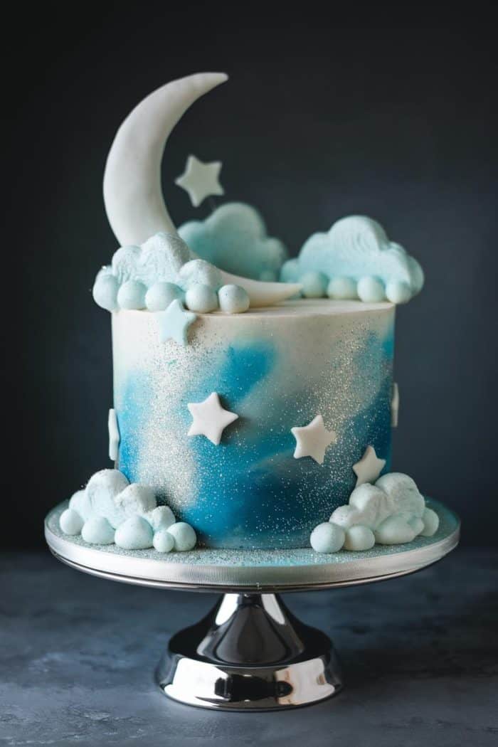 A whimsical cake decorated with a crescent moon, stars, and fluffy clouds. The cake is painted in shades of blue and white to resemble a night sky, and it sits on a shiny silver stand.