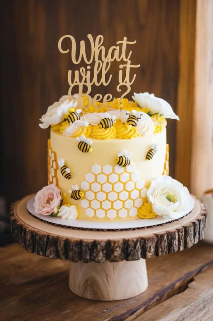 A yellow and white tiered cake with honeycomb patterns and small bee decorations. A wooden topper reads "What will it bee?" The cake is placed on a rustic wooden stand and adorned with white and pink flowers.