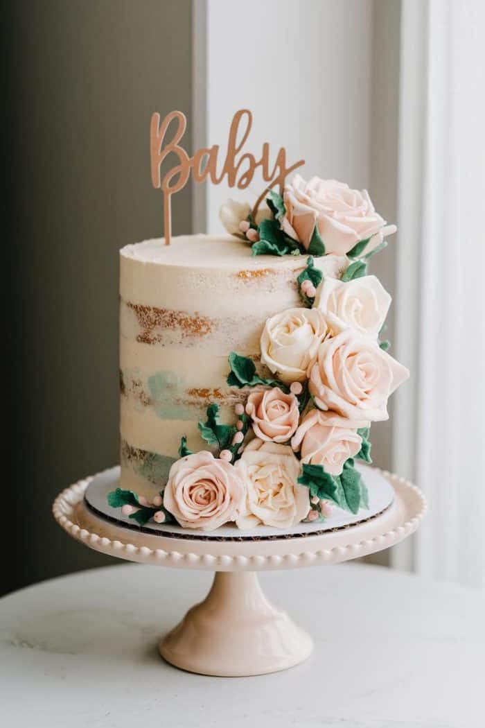 A tall, semi-naked cake on a pedestal stand, adorned with pastel pink and cream roses cascading down the side. The cake topper reads "Baby" in cursive lettering.
