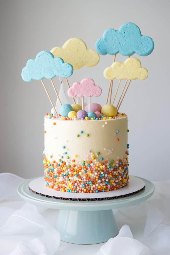 A colorful cake with a white frosting base, decorated with multicolored sprinkles and pastel miniature eggs. Cloud-shaped cookies on sticks are arranged on top. The cake is placed on a light blue cake stand against a neutral background.