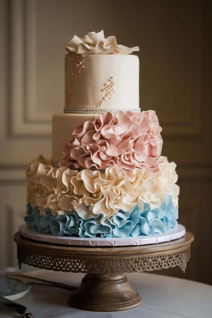 A three-tiered cake with elegant ruffled icing in shades of cream, pink, and blue stands on an ornate metal cake stand. The top tier is adorned with small floral details and pearl-like accents, creating a sophisticated and artistic look.