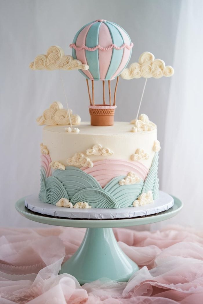 A pastel-themed cake with a hot air balloon and cloud decorations. The hot air balloon is perched on top, with pink and blue layers on the cake, accompanied by swirls and shaped clouds. It sits on a pastel green cake stand.