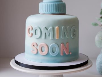 A blue and pastel-colored cake shaped like a baby bottle, with the words "COMING SOON" in pink and blue letters, sits on a white cake stand. The background is softly blurred with hints of greenery.