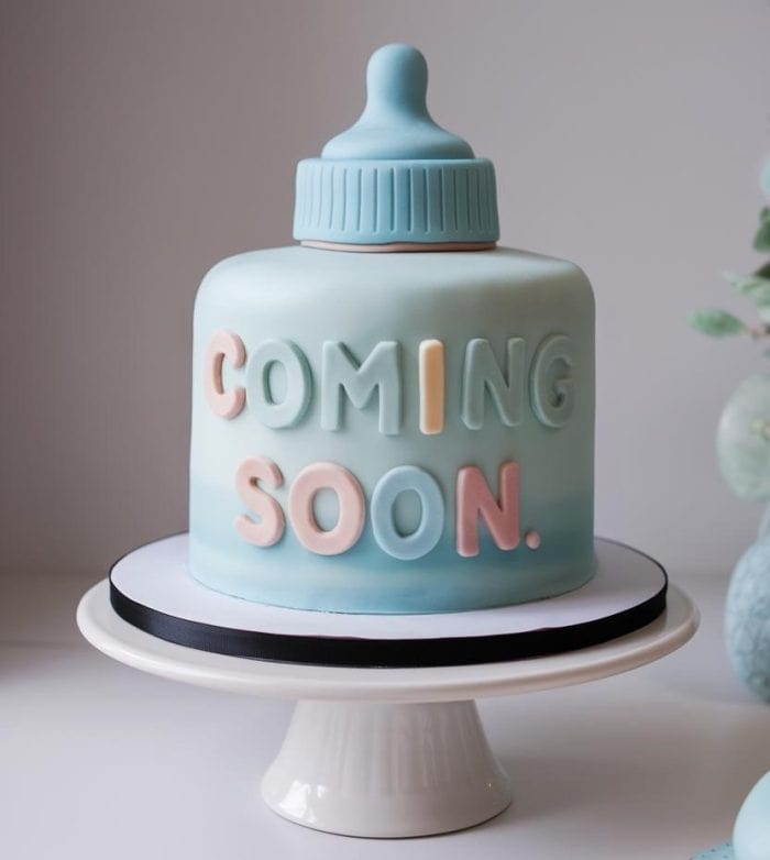 A blue and pastel-colored cake shaped like a baby bottle, with the words "COMING SOON" in pink and blue letters, sits on a white cake stand. The background is softly blurred with hints of greenery.