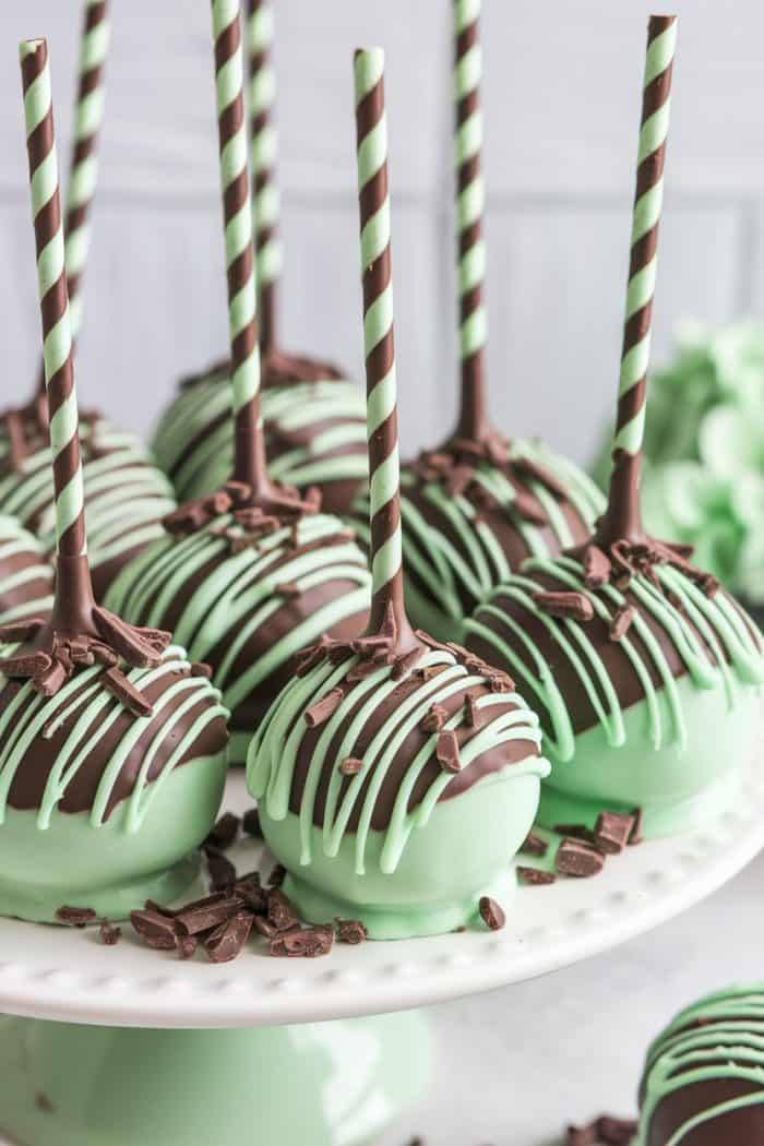 Close-up of mint chocolate cake pops on a white cake stand. The cake pops are coated in green and dark chocolate, with striped green and brown sticks, drizzled with chocolate, and sprinkled with small chocolate pieces.