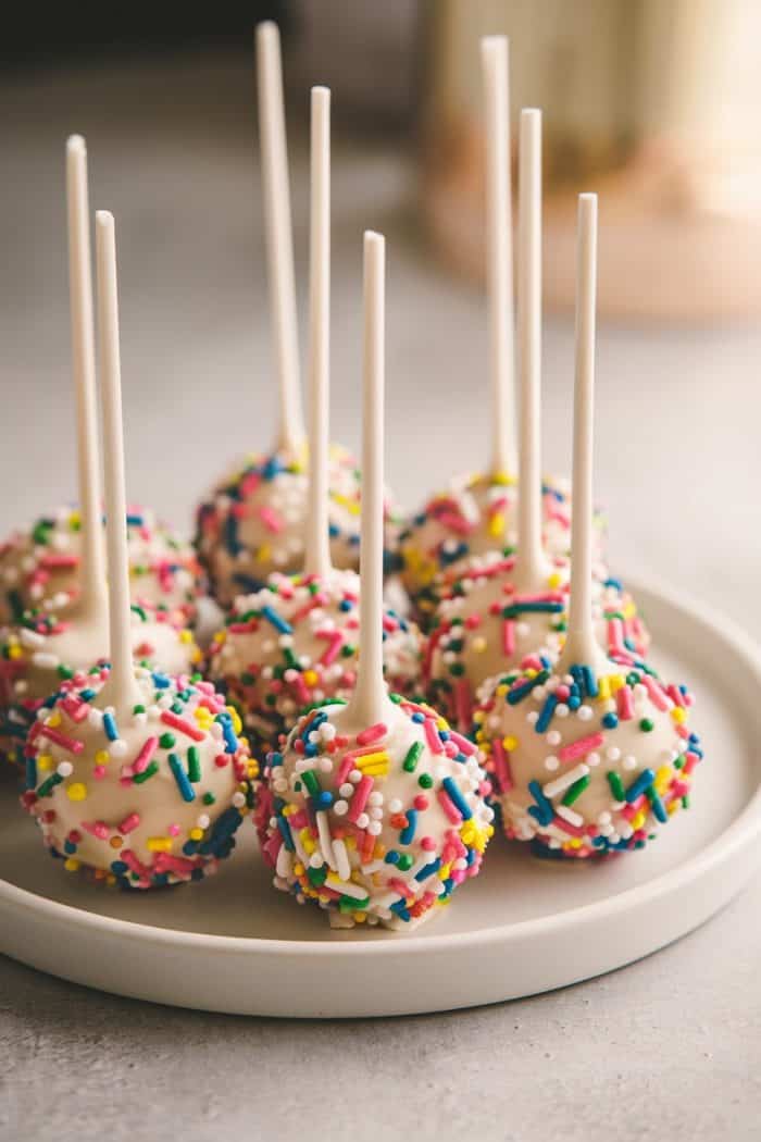 A plate of colorful cake pops with white icing and multicolored sprinkles. Each cake pop is on a white stick. They are arranged neatly on a smooth, round white plate.