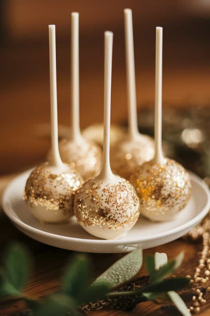 Five cake pops are elegantly displayed in a white dish. Each has a glossy white coating and is sprinkled with golden glitter, set against a blurred, warm-toned background.