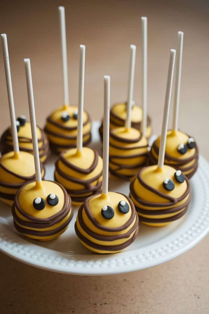 Bee-themed cake pops with yellow icing and chocolate stripes arranged on a white plate. Each pop has cute eyes and is topped with a stick.