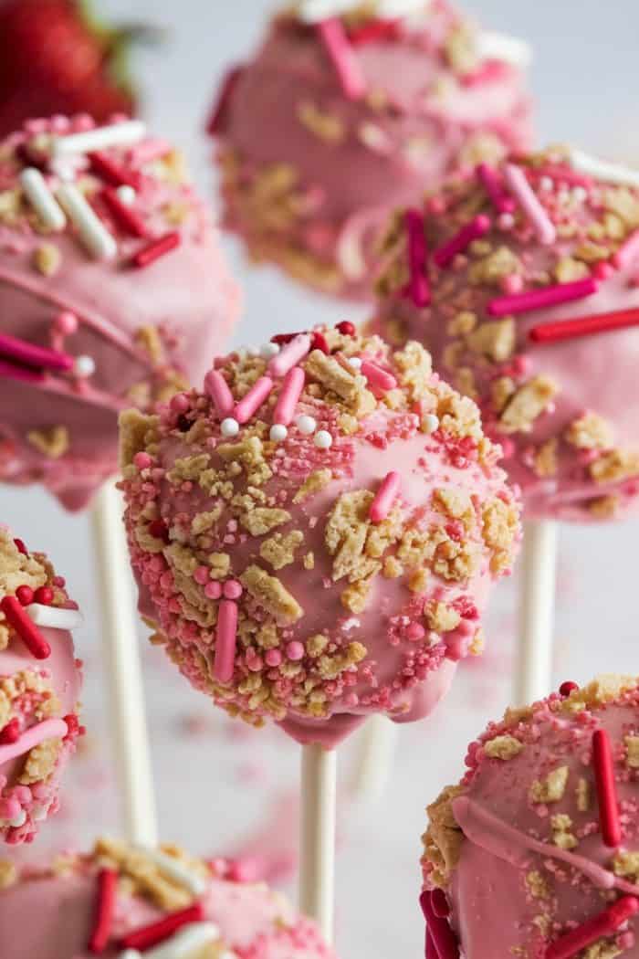 Close-up of pink cake pops with sprinkles and crushed cookie toppings on sticks. The cake pops are decorated with pink, white, and red sprinkles, and are set against a blurred light background.