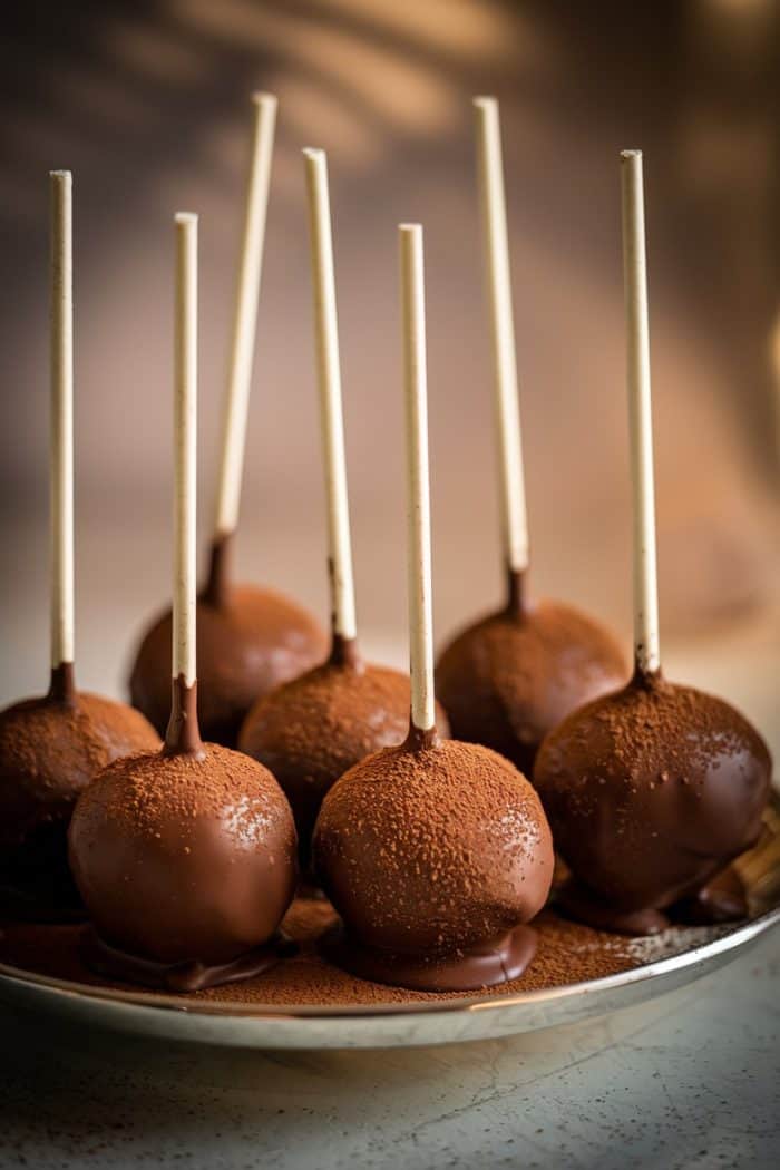 A plate of seven chocolate cake pops with wooden sticks. Each pop is dipped in chocolate and dusted with cocoa powder, creating a glossy and textured appearance. The background is softly blurred.