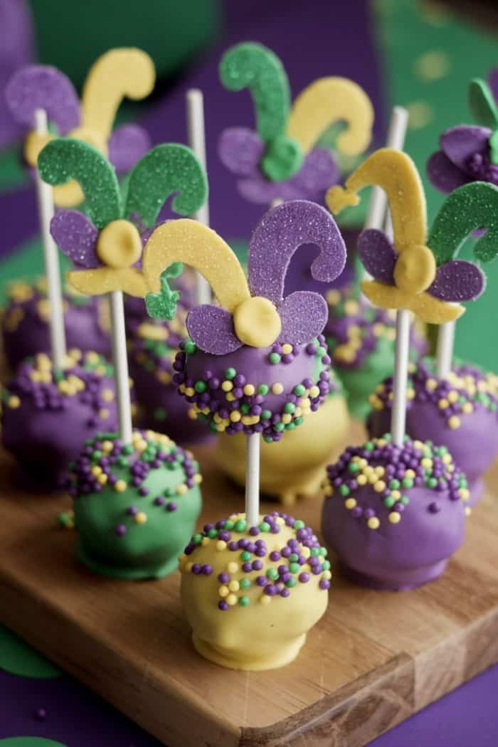 Festive cake pops decorated in Mardi Gras colors—purple, green, and yellow—topped with glittery, jester hat-shaped toppers, displayed on a wooden board.