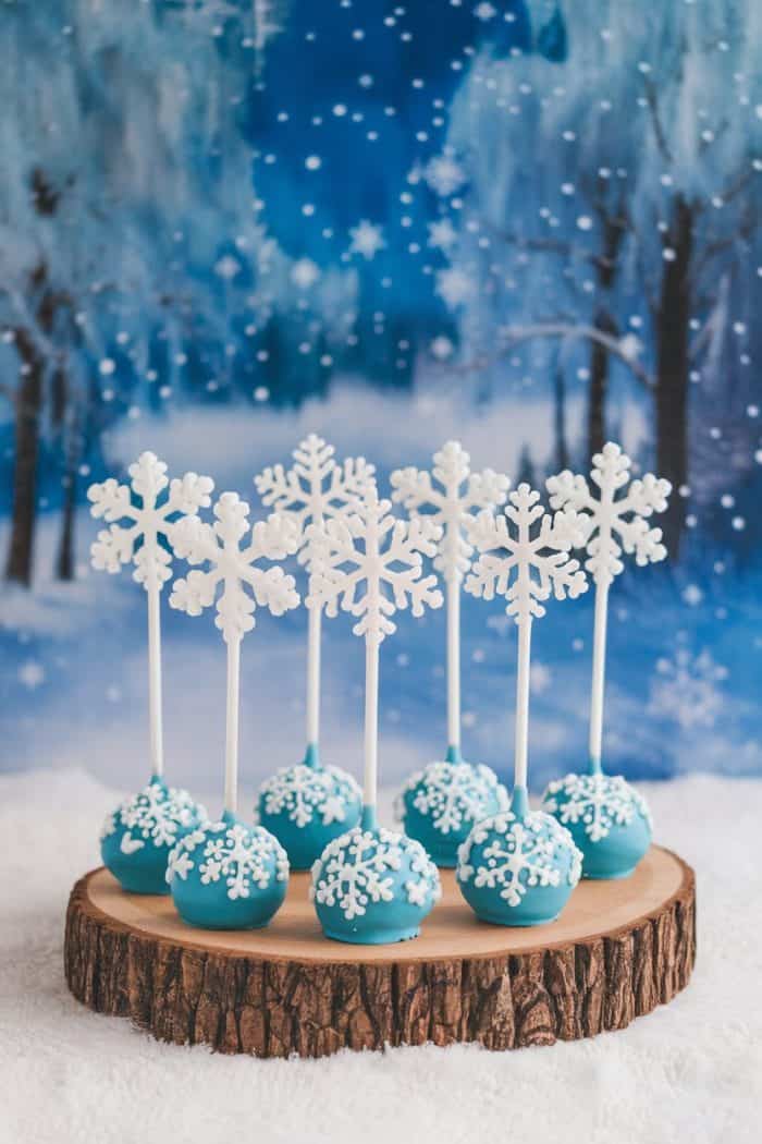 Eight blue cake pops with white snowflake decorations, topped with large snowflake toppers, are displayed on a wooden platter. The background features a snowy, wintry scene with trees and a midnight blue sky.