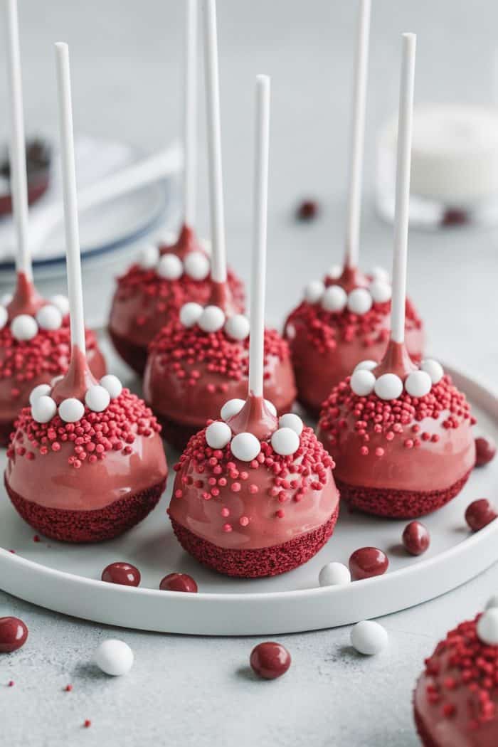 Red velvet cake pops are displayed on a white plate. Each pop is decorated with small red sugar sprinkles and white candy balls on top. The background features blurred elements of another plate and some scattered candy.