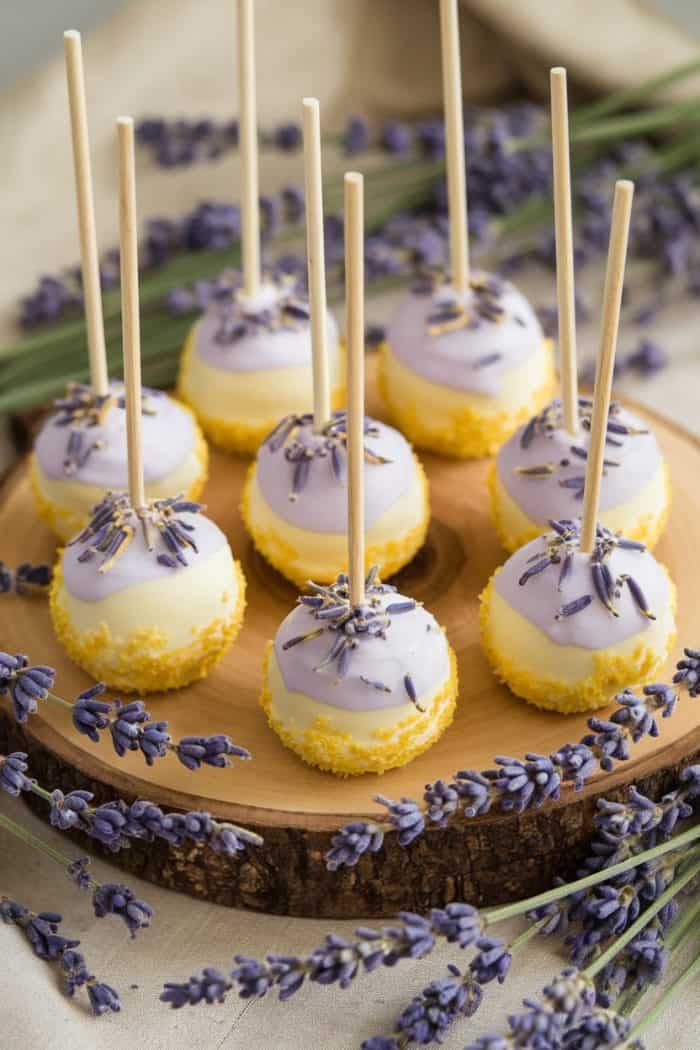 Cake pops with purple and white icing are displayed on a wooden platter, garnished with lavender sprigs. Each pop is topped with dried lavender, creating a visually appealing and aromatic dessert presentation.