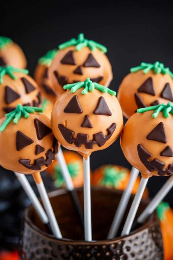 Halloween-themed cake pops shaped like jack-o'-lanterns with orange coating and chocolate faces. Green icing tops represent stems. They are displayed in a textured, dark bowl against a black background.