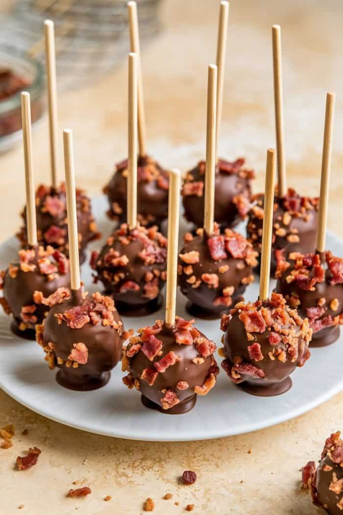 Chocolate cake pops topped with chopped bacon are arranged on a white plate. Each cake pop is on a wooden stick, creating a savory and sweet presentation. The background is a neutral, light-colored surface.
