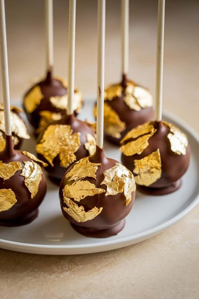 Chocolate cake pops coated in gold leaf are arranged on a white plate. Each pop is mounted on a white stick, giving a luxurious and elegant appearance.