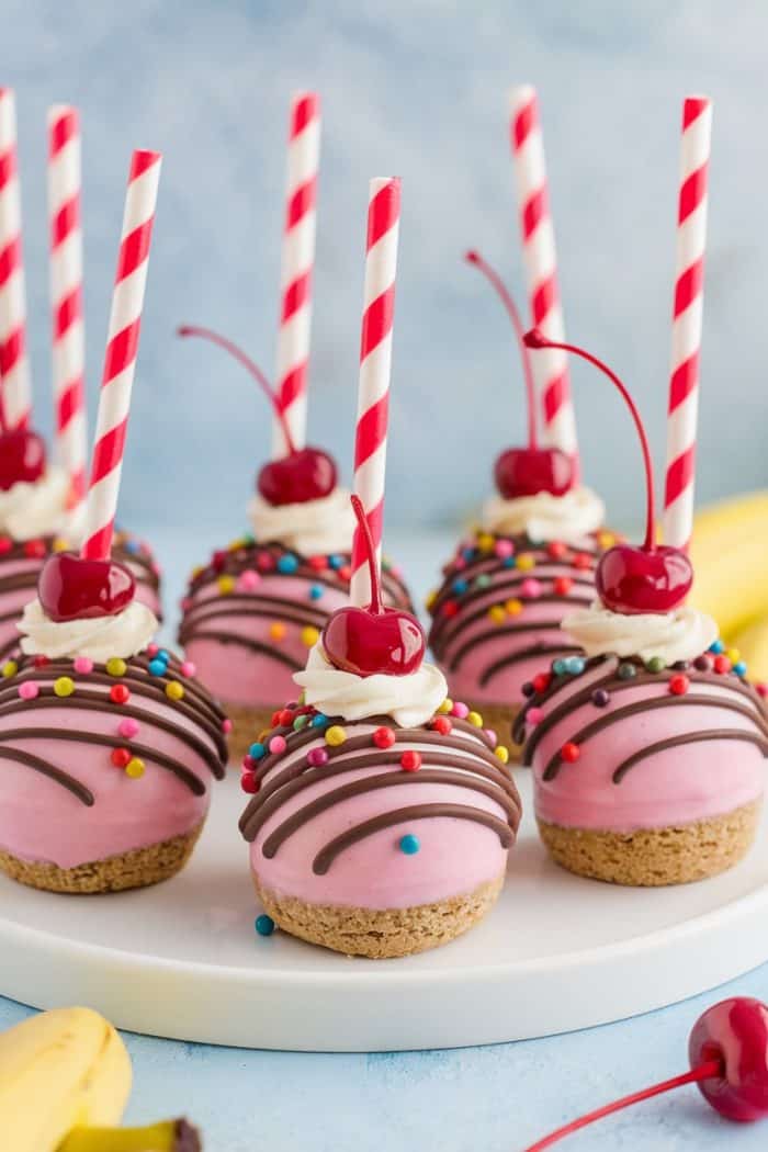 Pink dessert balls topped with chocolate drizzle, colorful sprinkles, whipped cream, and cherries. Each has a red and white striped straw. They are arranged on a white plate with blurred bananas in the background.