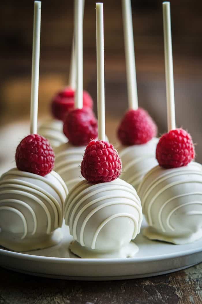 White chocolate cake pops with intricate swirl designs, topped with fresh raspberries, are arranged on a white plate. They each have a stick for holding, and the presentation is elegant and inviting.