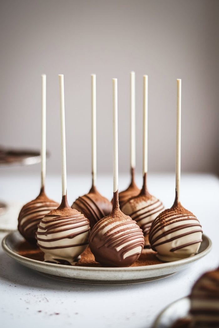A plate of chocolate cake pops with white sticks, drizzled with milk and white chocolate, neatly arranged on a white surface. The background is softly blurred.