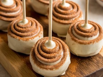 Cake pops with swirled frosting and cinnamon sprinkle on sticks, arranged on a wooden board. A bowl of cinnamon and a few sticks are visible in the background.