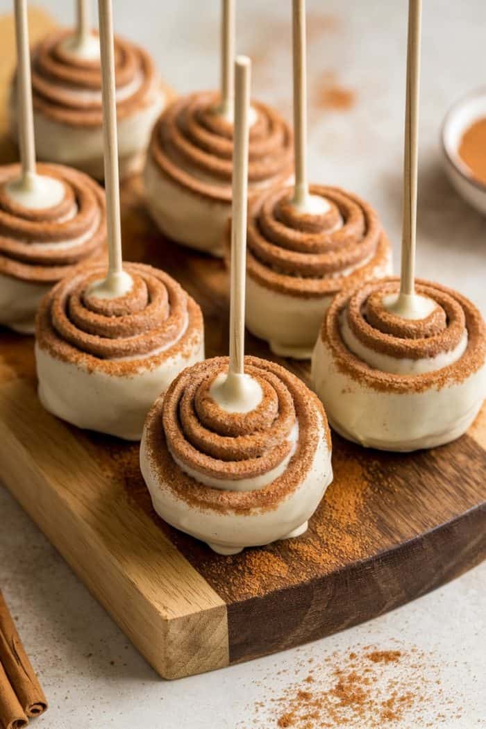 Cake pops with swirled frosting and cinnamon sprinkle on sticks, arranged on a wooden board. A bowl of cinnamon and a few sticks are visible in the background.