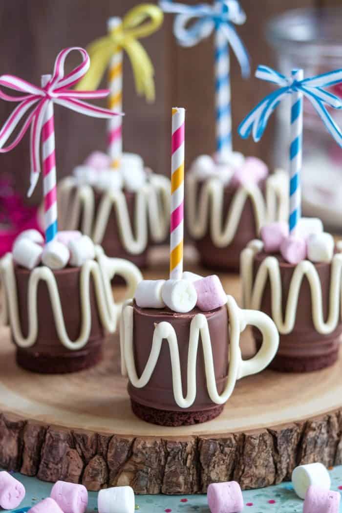 Chocolate cups designed to look like mini mugs, topped with marshmallows and decorated with white icing. Each has a colorful striped straw and ribbon, arranged on a wooden board. Perfect for a festive or whimsical dessert display.
